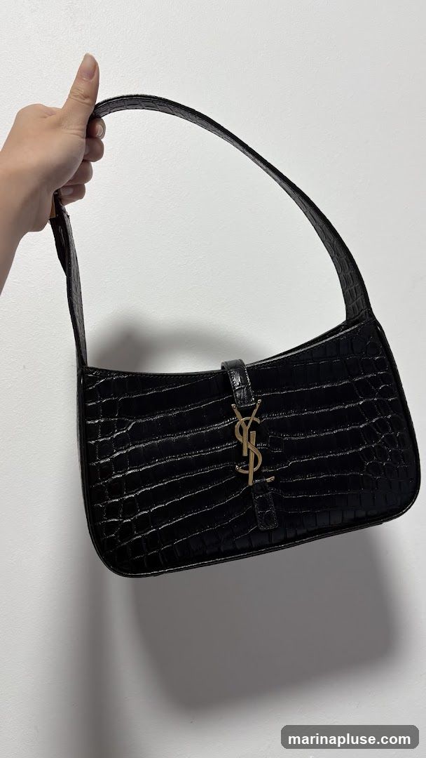 A black shoulder bag with a crocodile embossed texture is held by a hand against a white background. The bag features a gold-toned YSL logo on the front flap.