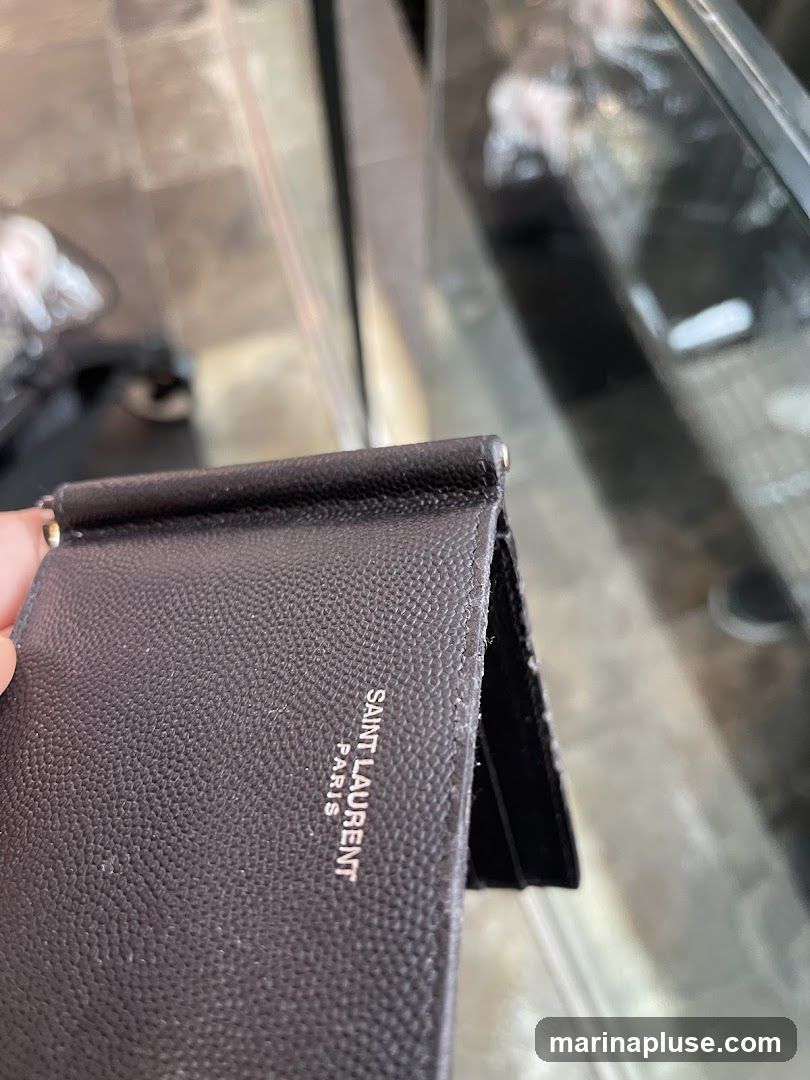 A close-up shot of a black Saint Laurent money clip wallet. The wallet is made of textured leather and features the brand's logo embossed in silver on the interior. The background is blurred, showing a reflective surface and what appears to be a store display.