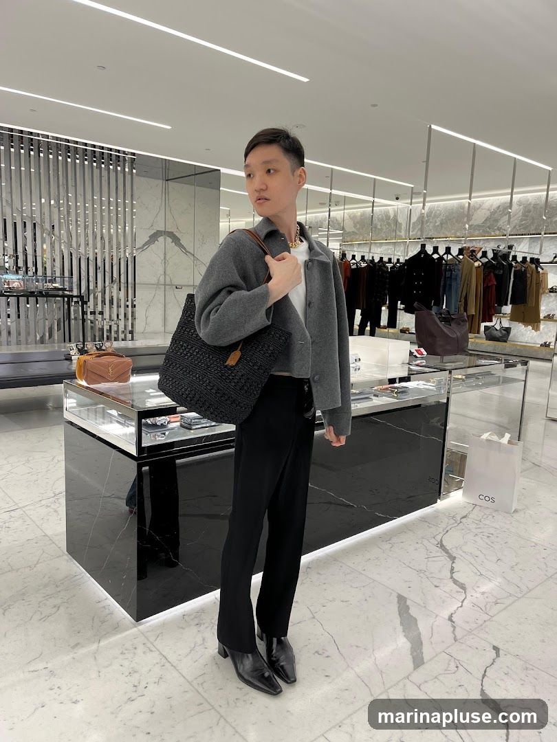 A person stands in a well-lit store, holding a black woven tote bag. They are wearing a grey cropped jacket over a white t-shirt, black pants, and black heeled boots. The background shows racks of clothing, display cases with merchandise, and a white shopping bag with the COS logo.