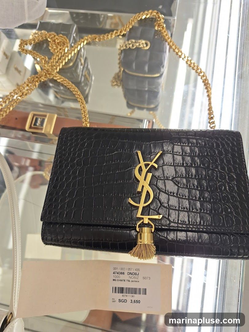 A black crocodile-embossed clutch bag with a gold YSL logo and tassel detail. The bag has a gold chain strap. A price tag is visible below the bag, showing the item code, description, and price.