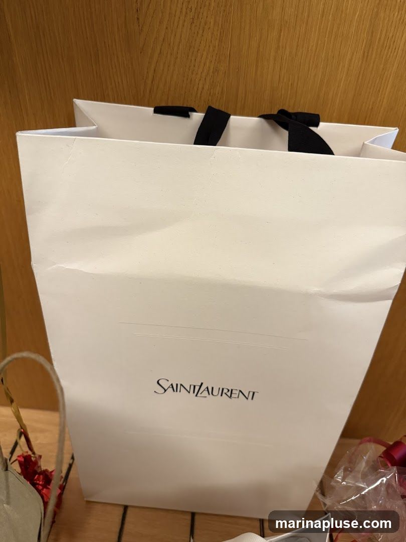 A white Saint Laurent shopping bag with black ribbon handles is shown. The Saint Laurent logo is printed in black on the front of the bag. There are also glimpses of other items, including a small basket with red flowers and a gift-wrapped item with a pink ribbon. The background appears to be a wooden surface.
