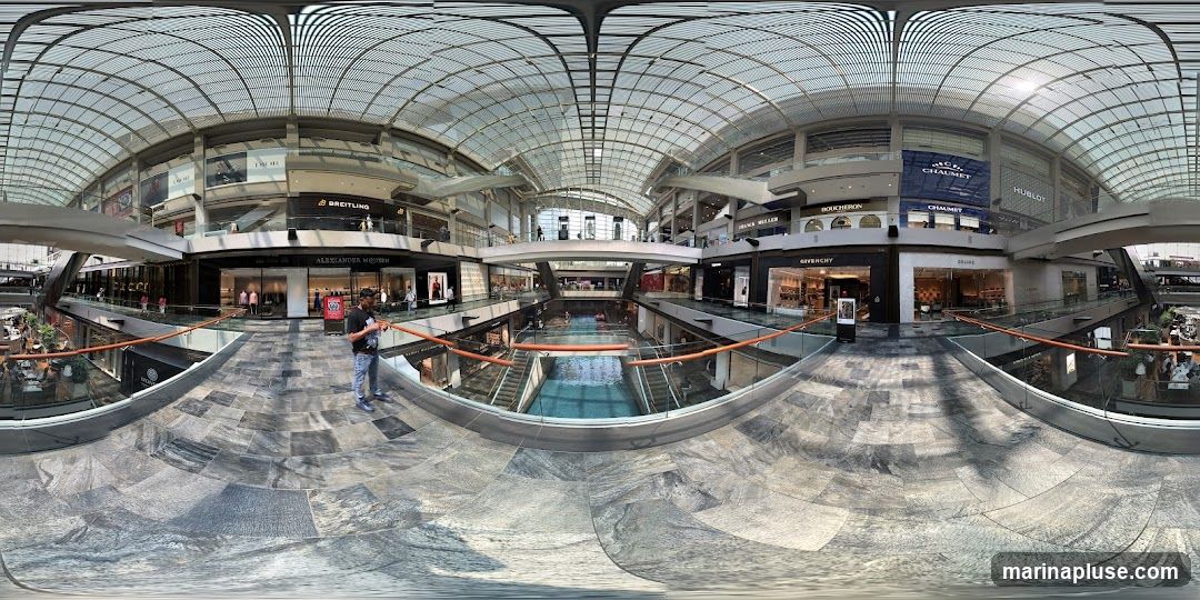 A panoramic view of a large, modern indoor shopping mall. The mall features multiple levels with various luxury brand stores visible, including Dior, Breitling, Alexander McQueen, Boucheron, Chaumet, Hublot, Givenchy, and Celine. A central canal or water feature runs through the lower level, with escalators and walkways connecting different sections. The architecture includes a distinctive glass roof structure. People are seen browsing in stores and walking around the mall.