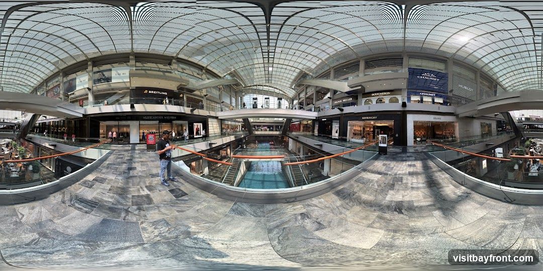A panoramic view of a large, modern indoor shopping mall. The mall features multiple levels with various luxury brand stores visible, including Dior, Breitling, Alexander McQueen, Boucheron, Chaumet, Hublot, Givenchy, and Celine. A central canal or water feature runs through the lower level, with escalators and walkways connecting different sections. The architecture includes a distinctive glass roof structure. People are seen browsing in stores and walking around the mall.