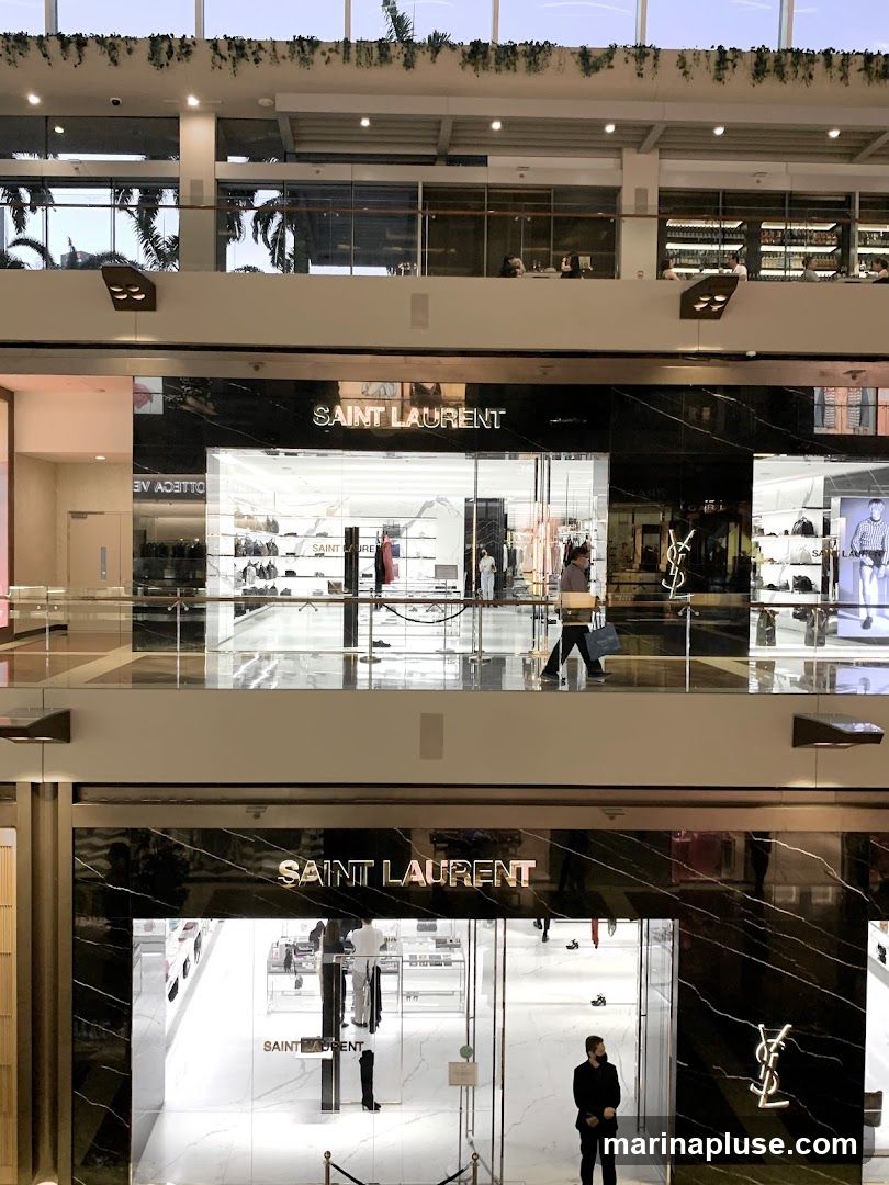 A view of two Saint Laurent store entrances on different levels of a mall. The storefronts feature black marble facades with the Saint Laurent logo and name prominently displayed. Inside, merchandise such as handbags and clothing are visible on display shelves and racks.