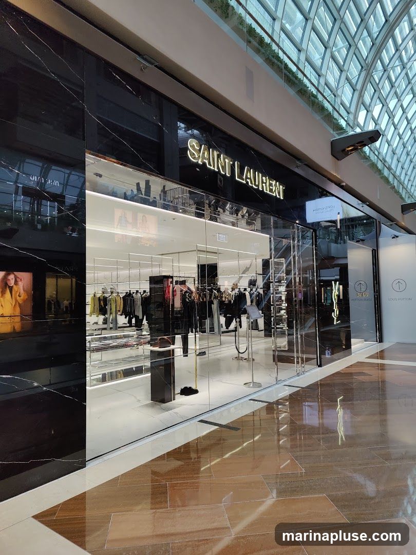 The exterior of a Saint Laurent store with a large glass facade. The store's name is prominently displayed in gold lettering above the entrance. Inside, racks of clothing and display shelves are visible. To the right, a Louis Vuitton store is also visible.
