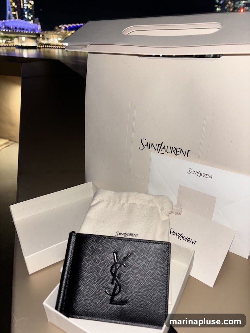 A black Saint Laurent wallet with the YSL logo is displayed in its white box. Next to it is a white dust bag and a Saint Laurent shopping bag. In the background, a city skyline at night is visible.