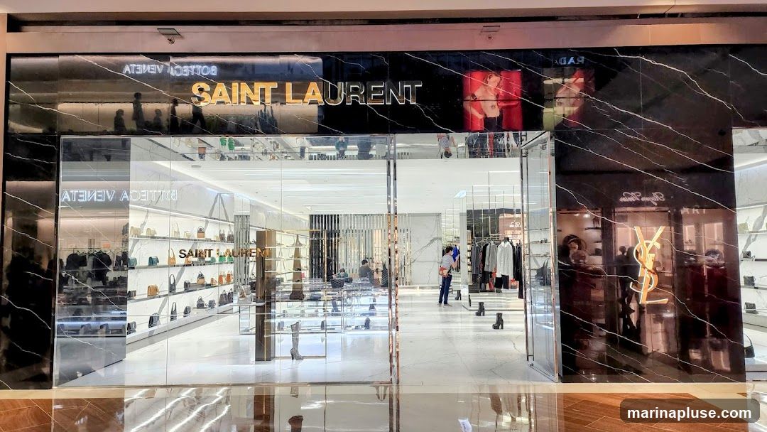 The exterior of a Saint Laurent store with a black marble facade. The store name "SAINT LAURENT" is displayed in large gold lettering above the entrance. The interior is visible through large glass windows, showing displays of handbags and clothing. To the left, "BOTTEGA VENETA" is visible in reflection. To the right, the Yves Saint Laurent logo is displayed in gold.
