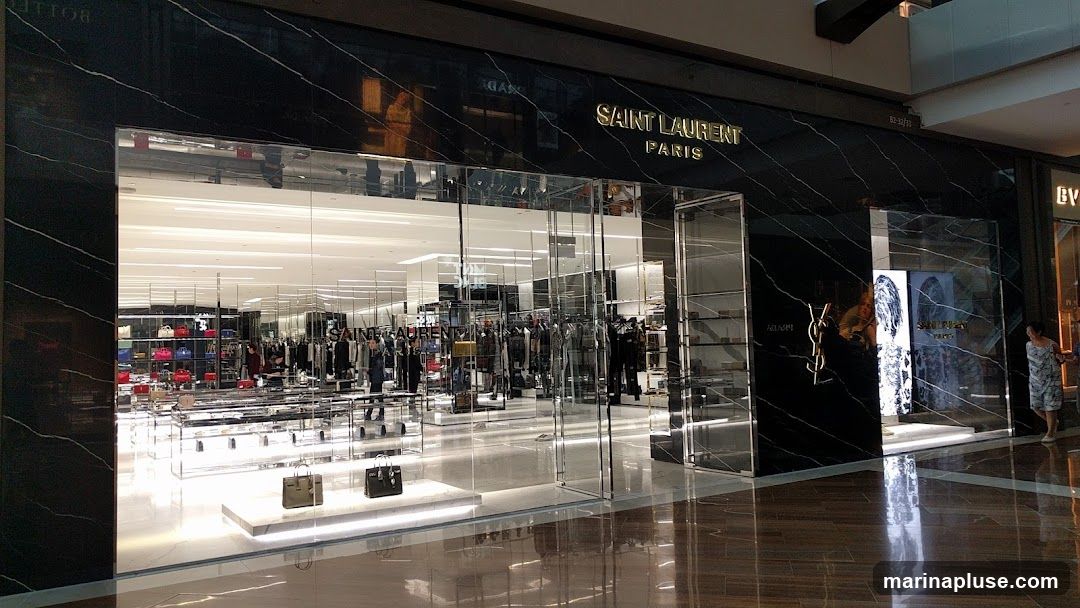 The exterior of a Saint Laurent Paris store. The facade is made of black marble with gold lettering that reads "SAINT LAURENT PARIS". The large glass windows reveal a brightly lit interior with clothing racks and display shelves filled with handbags. Two handbags are displayed on a pedestal in front of the store.