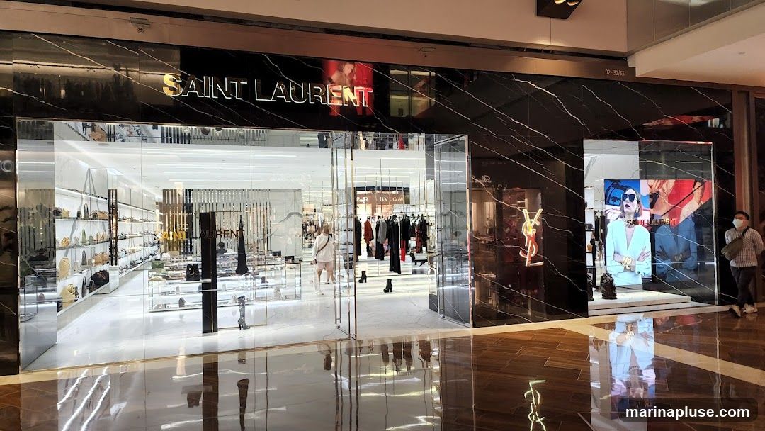 The exterior of a Saint Laurent store with a black marble facade. The store name is displayed in gold lettering above the entrance. The interior is visible through large glass windows, showing displays of handbags, clothing, and shoes. A digital screen displays an advertisement with a model wearing sunglasses.