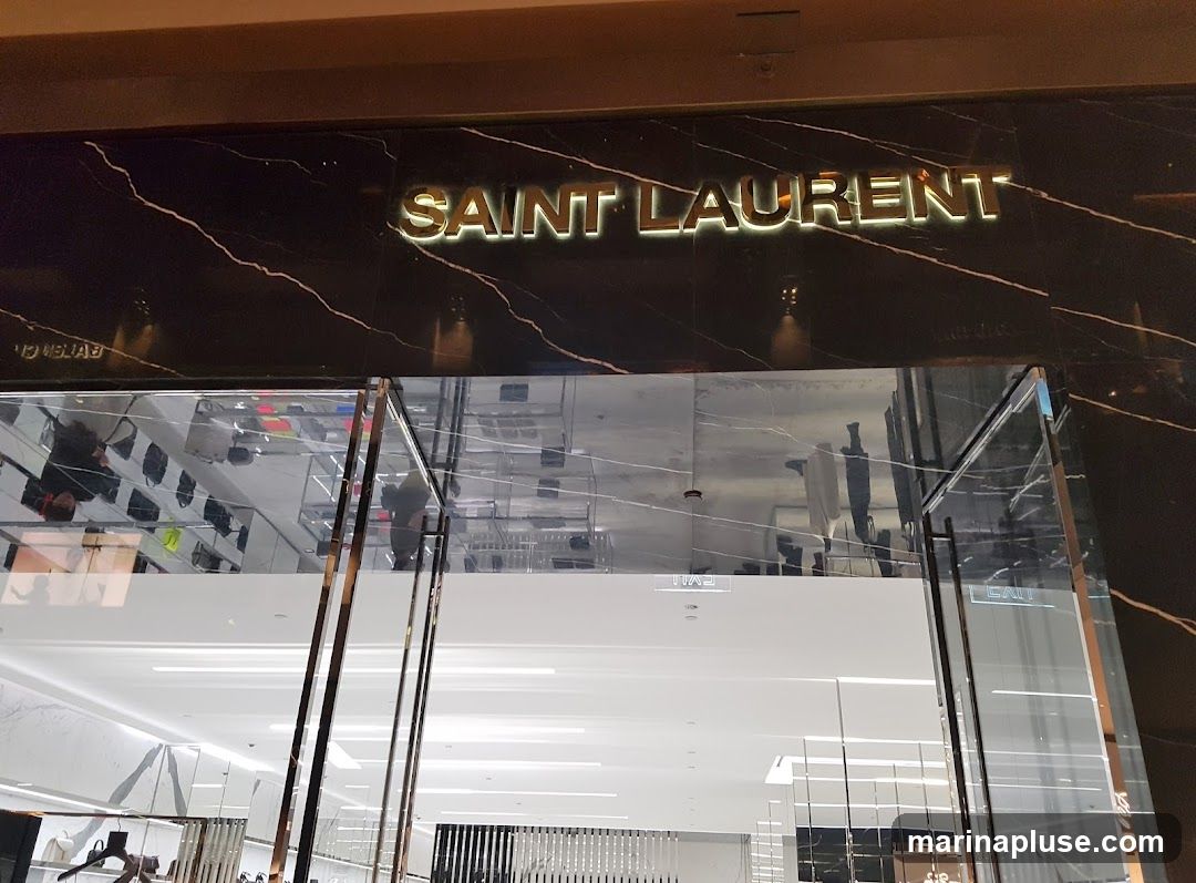 The exterior of a Saint Laurent store with the brand name displayed in gold lettering on a black marble facade. The entrance features large glass doors and windows reflecting the interior of the store, which appears to be a high-end boutique with displays of merchandise.