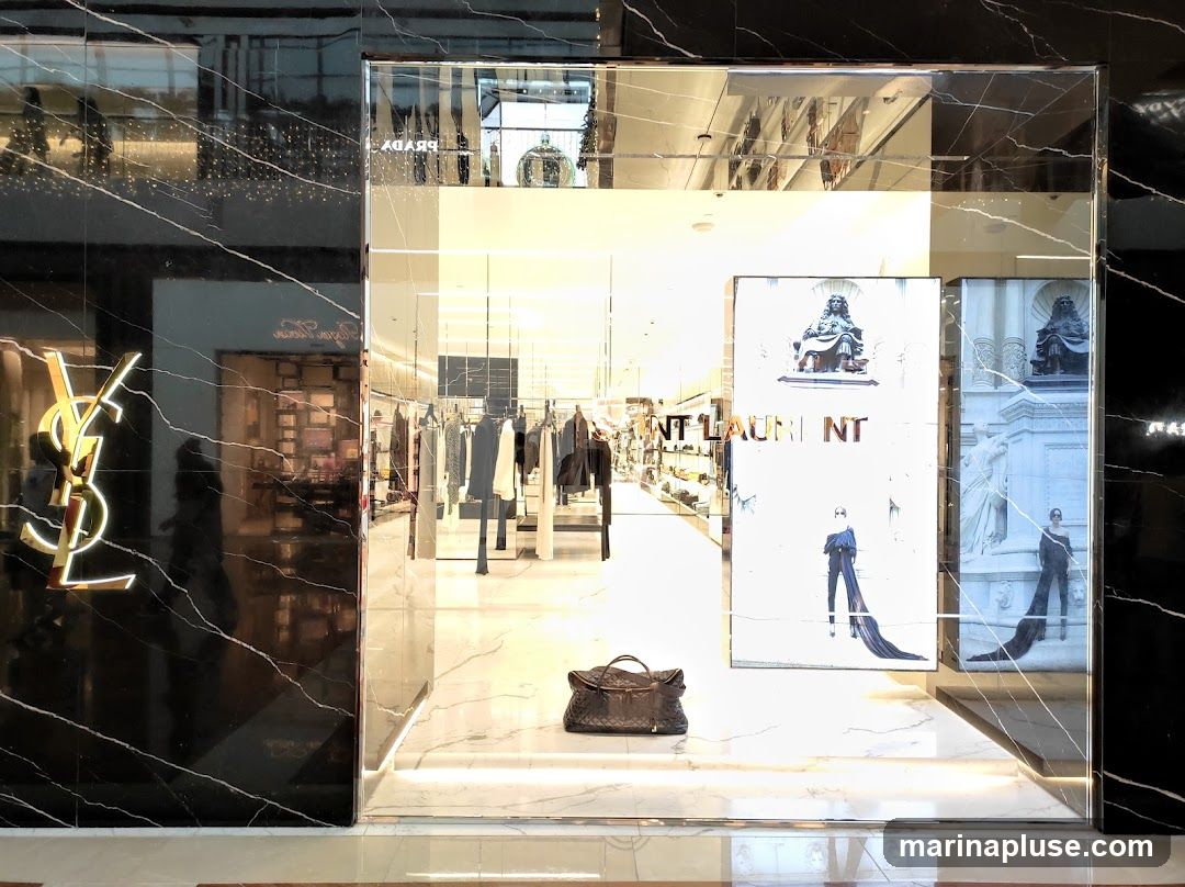 A luxury fashion store's exterior features a prominent gold YSL logo on a black marble facade. The large display window showcases a collection of clothing on racks, a dark quilted duffel bag on the floor, and large advertisements featuring models and statues. The interior is brightly lit, revealing more merchandise and displays.