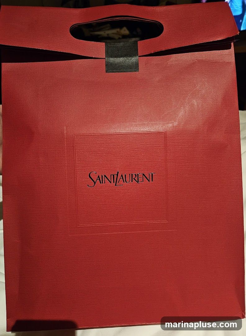 A red textured shopping bag with the Saint Laurent logo embossed in the center. The bag has a black strap detail and cut-out handles.