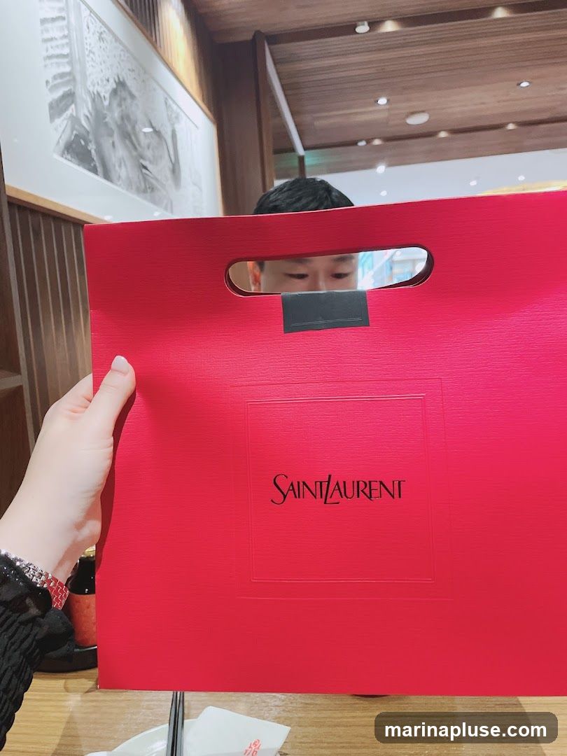 A person holds a bright red Saint Laurent shopping bag with the brand's logo embossed in the center. The bag has a cutout handle. In the background, a person's face is visible peeking through the handle, and a framed artwork hangs on the wall. The setting appears to be indoors, possibly a restaurant or cafe, with wooden paneling and ceiling lights visible.