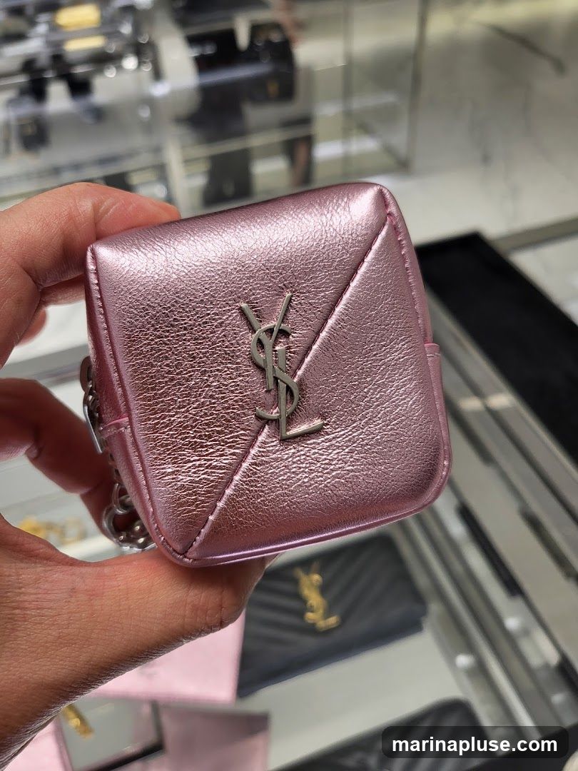 A hand holds a small, pink, metallic pouch with a quilted pattern and the YSL logo in silver. In the background, other luxury items like wallets and bags are displayed in a store setting.