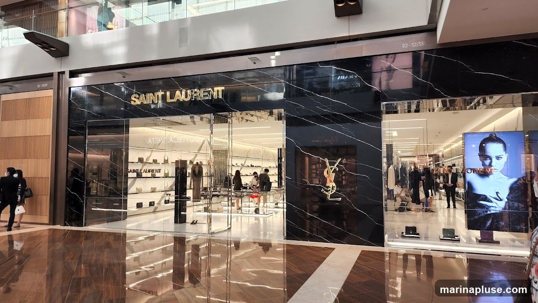 The exterior of a Saint Laurent store with a black marble facade. The store name is displayed in gold lettering above the entrance. Inside, merchandise such as handbags and clothing are visible on display shelves and racks. A large advertisement featuring a woman is displayed on the right side of the storefront.