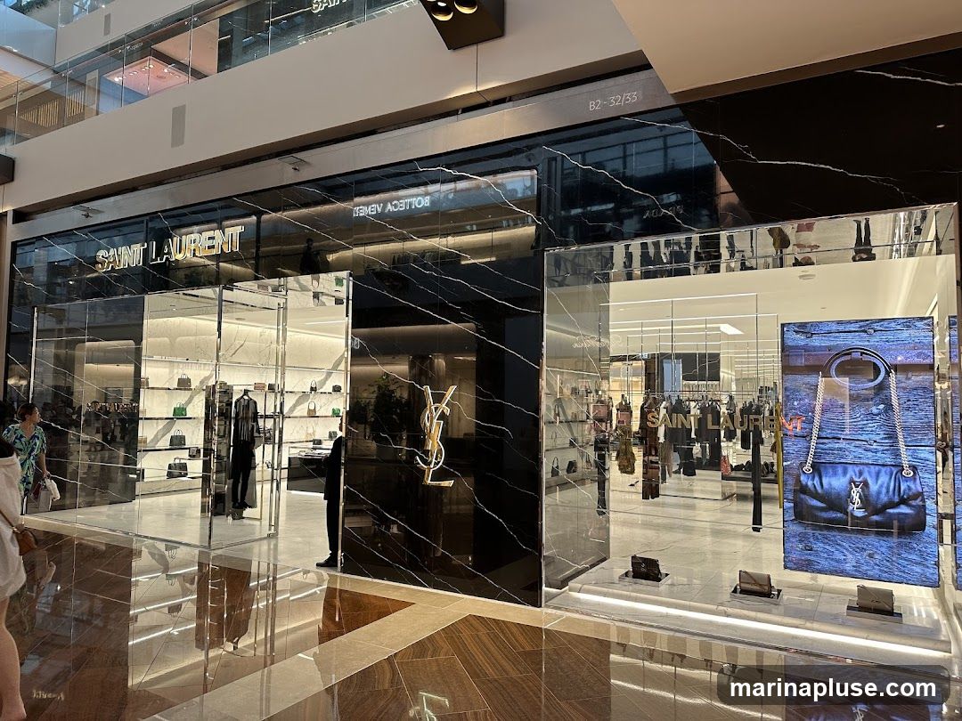 The exterior of a Saint Laurent store with a black marble facade and large glass windows displaying handbags and clothing. The Saint Laurent logo is visible on the facade.