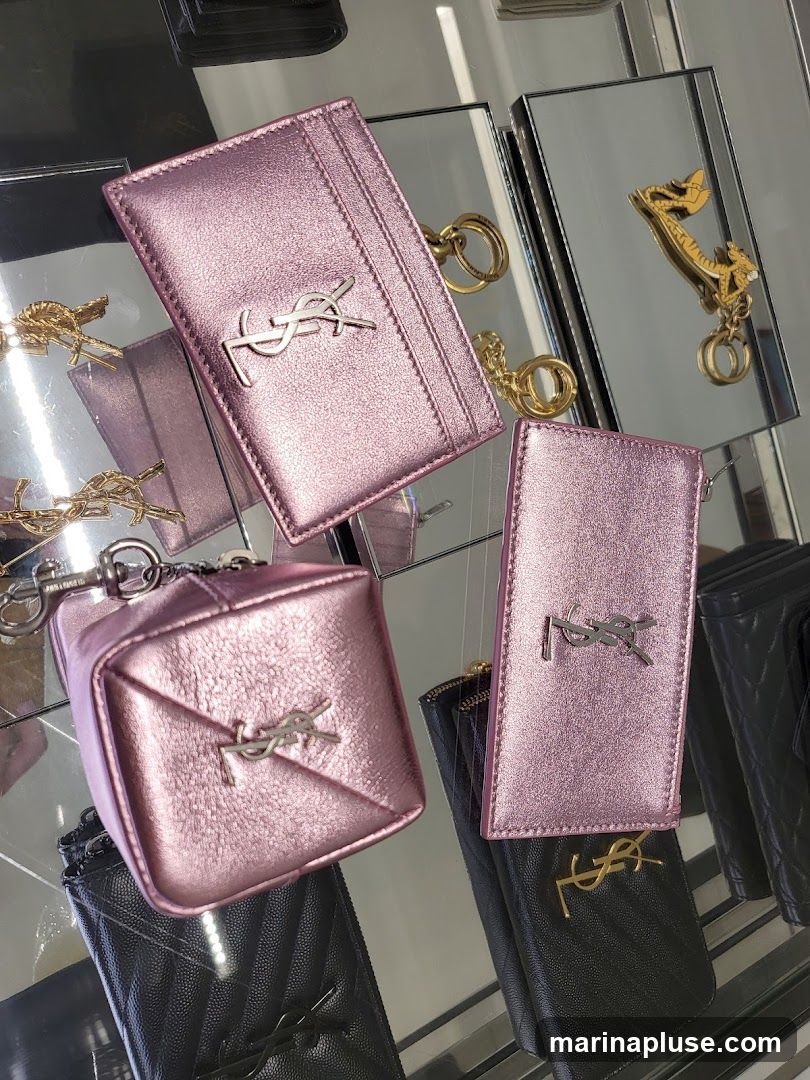 A display of pink metallic accessories, including a cardholder with a keychain, a small pouch, and another cardholder, all featuring the YSL logo. Black quilted wallets with the YSL logo are visible underneath. The items are displayed on a reflective surface, possibly a glass shelf, with mirrors reflecting the items and a tiger-shaped keychain.