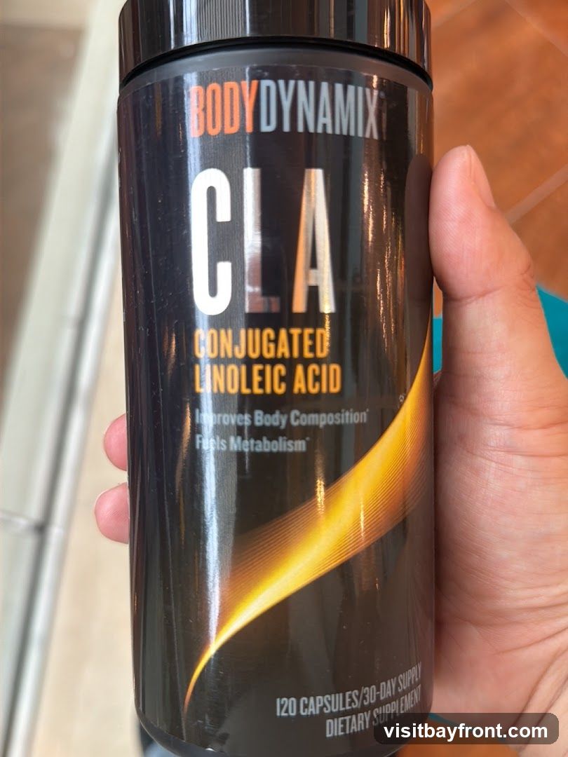 A hand holds a black bottle of Body Dynamix CLA (Conjugated Linoleic Acid) supplement. The label states it improves body composition and fuels metabolism. It contains 120 capsules, a 30-day supply, and is a dietary supplement.
