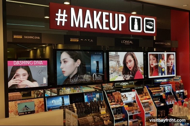 A brightly lit display of makeup products and advertisements from various brands including Dashing Diva, Kate, L'Oréal, and Maybelline. The prominent sign above reads "#MAKEUP" with icons for lipstick and an eye. Digital screens showcase models and product information. Shelves are stocked with numerous makeup items.