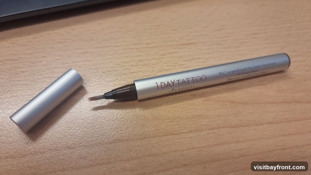 A silver eyeliner pen with a brown tip and cap is shown on a wooden surface. The pen has text printed on it that reads "1 DAY TATTOO -Procast-" and "We've perfected the basic functions of an eyeliner. The new standard eyeliner.".