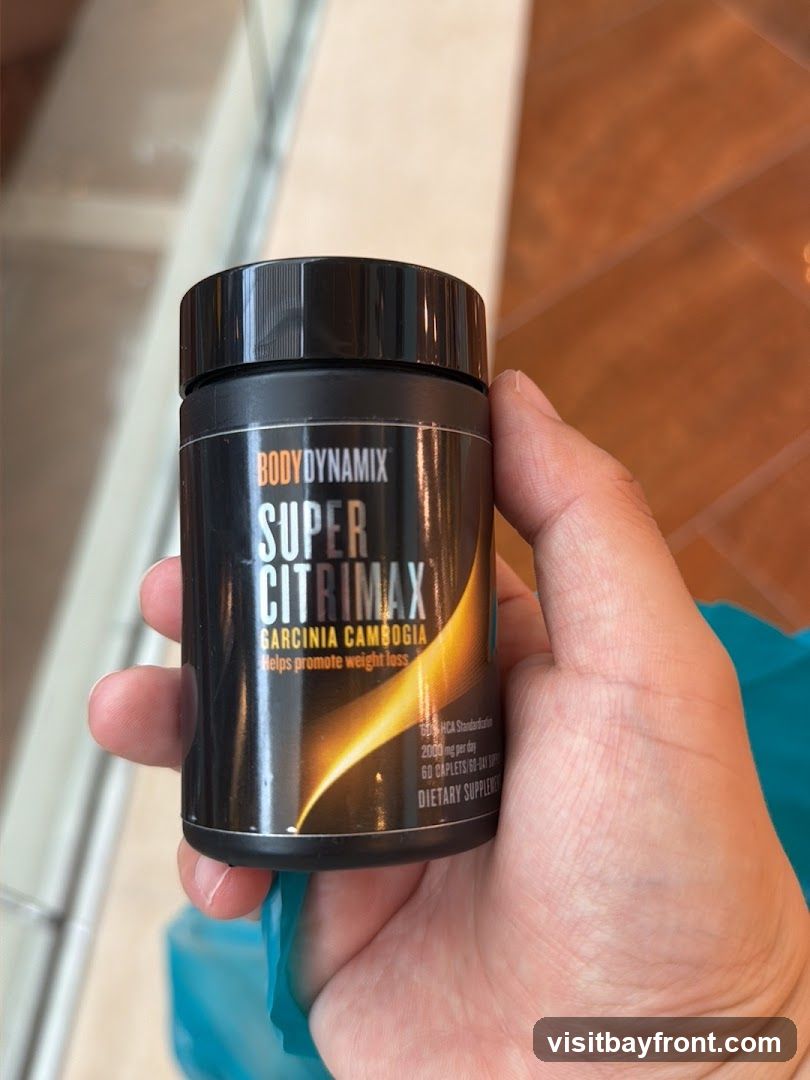 A hand holds a black bottle of Body Dynamix Super Citrimax, a dietary supplement containing Garcinia Cambogia, which claims to help promote weight loss. The label indicates it contains 2000 mg of HCA per day and 60 caplets.