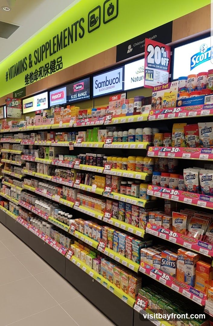 A wide shot of a brightly lit aisle in a store, filled with shelves stocked with various vitamins, supplements, and health products. The shelves are organized by brand and product type, with many items displayed in their original packaging. Promotional signs indicating discounts, such as '20% OFF' and '2 FOR $30', are visible throughout the aisle. Brands like Centrum, Swisse, Sambucol, and Scott's are prominently featured.