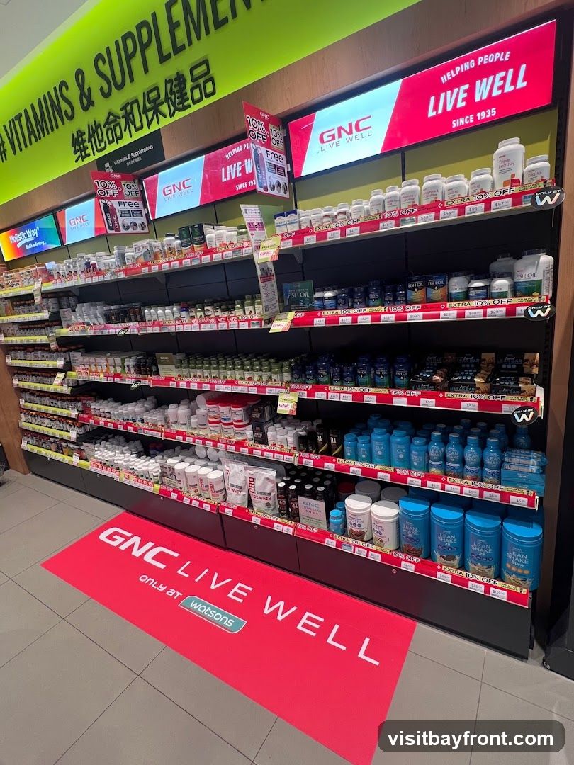 A retail display featuring shelves stocked with various vitamins, supplements, and health products. Prominent signage includes "# VITAMINS & SUPPLEMENTS" in large green lettering, and "GNC LIVE WELL" branding throughout. Red floor mats with "GNC LIVE WELL only at watsons" are visible. Several promotional signs indicate "10% OFF" and "EXTRA 10% OFF". Products include bottles of supplements, tubs of protein powder, and bottles of fish oil.