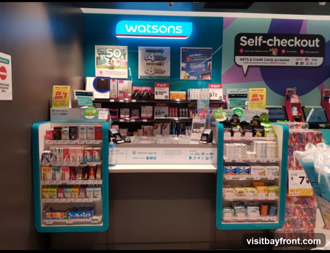 A Watsons store self-checkout area with various products displayed on shelves and promotional signage. The Watsons logo is visible. Products include health and beauty items, snacks, and personal care products. Promotions include