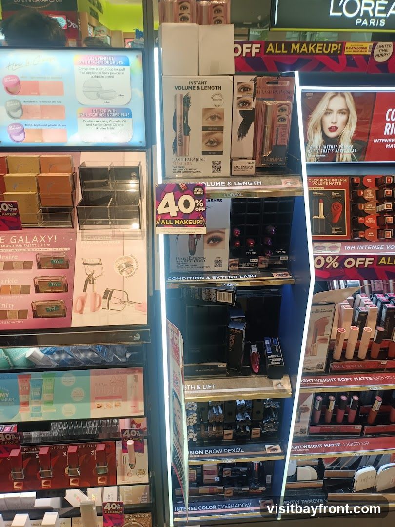 A brightly lit display of various makeup products, including mascaras, eyeshadow palettes, lipsticks, and brow pencils. Promotional signs indicate "40% OFF ALL MAKEUP!".