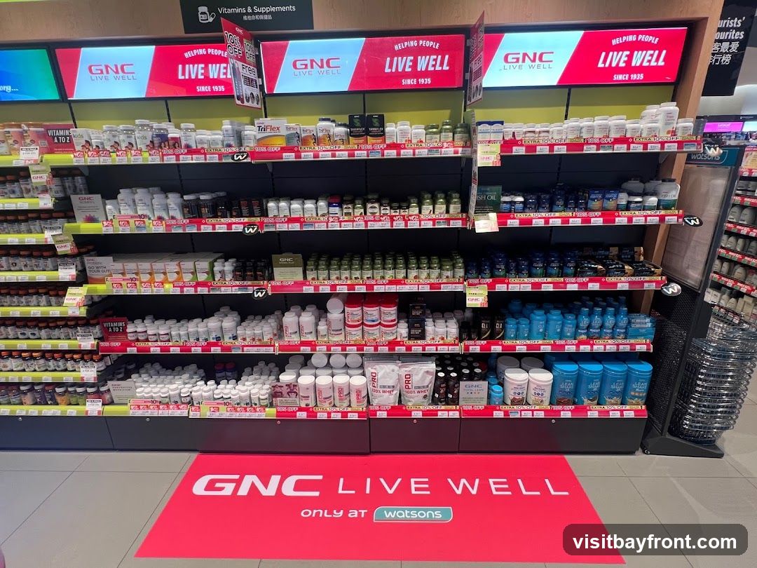 A retail store display featuring shelves stocked with various vitamins, supplements, and health products. The GNC brand is prominently displayed on signage and product packaging. A large red floor mat with the GNC Live Well logo and "only at Watsons" is visible in the foreground.