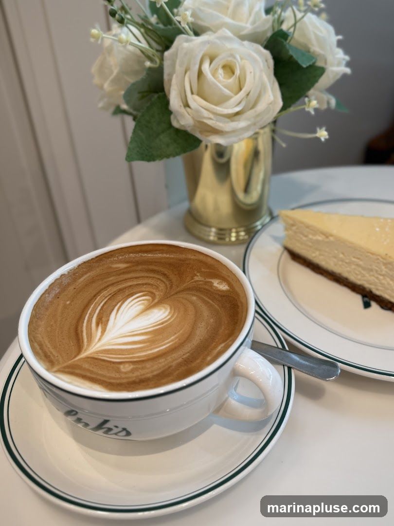 A close-up shot of a latte with intricate latte art in a white mug with green trim. Next to the mug is a slice of cheesecake on a white plate with green trim. A silver spoon rests on the plate. In the background, a vase of white roses adds a decorative touch.