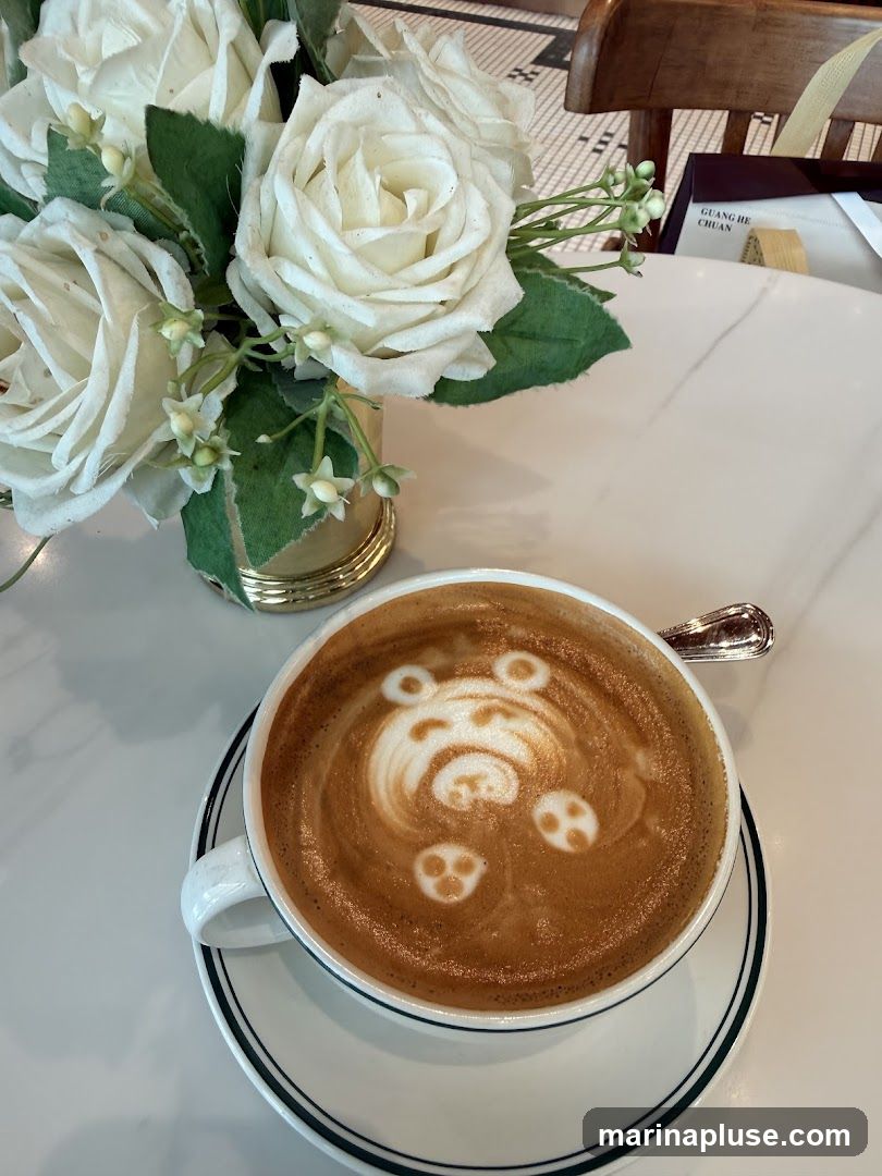 A white cup and saucer hold a latte with intricate foam art depicting a bear face. A bouquet of white roses with green leaves is arranged in a gold vase to the left of the cup. The table surface is white with a marble pattern. In the background, a wooden chair and a box with Chinese text are visible.