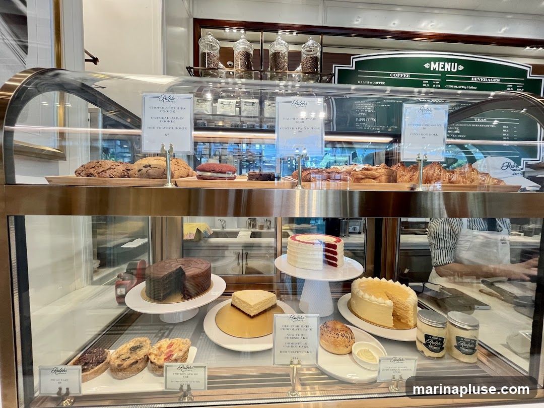 A display case filled with various desserts and baked goods, including cakes, cookies, croissants, and bagels. There are also jars of what appear to be spreads or toppings. A menu board is visible in the background.