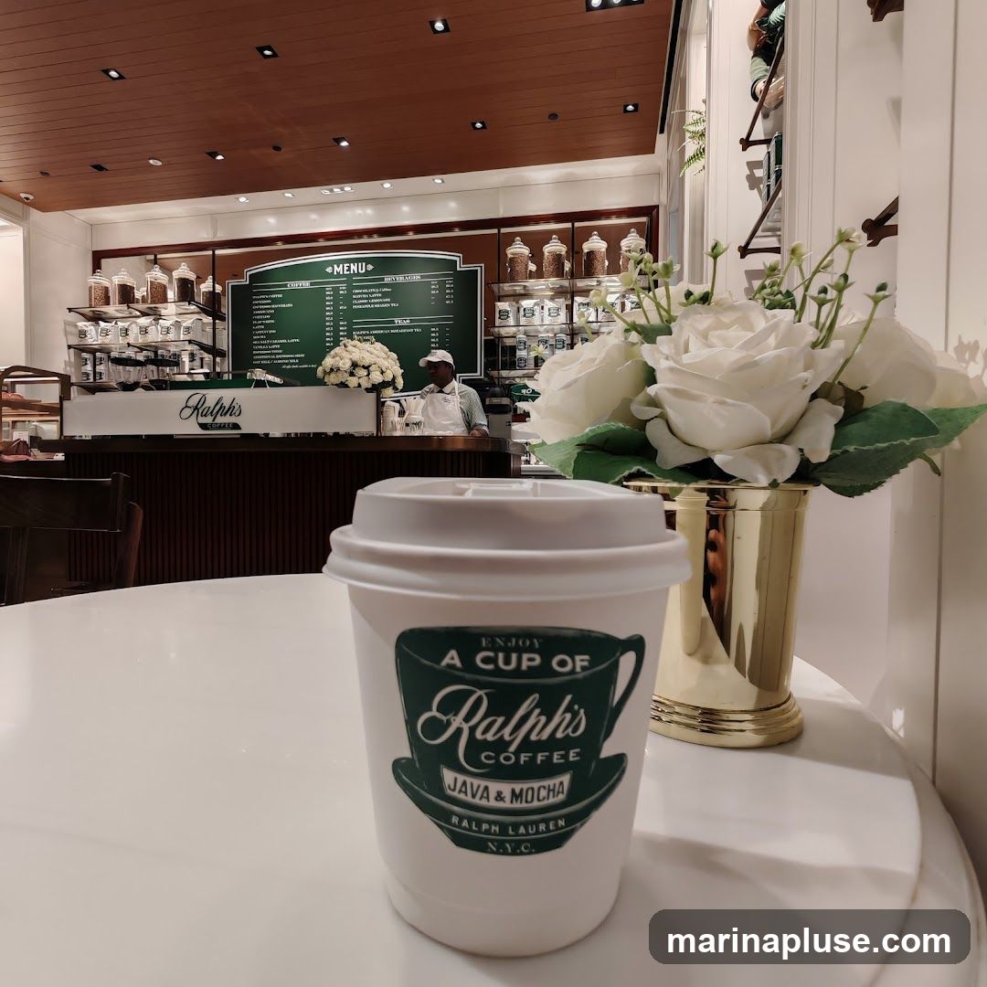 A white coffee cup with the Ralph's Coffee logo is in the foreground, placed on a white table. In the background, a coffee shop counter is visible with a menu board, jars of coffee beans, and a barista. A vase of white roses is to the right of the cup.