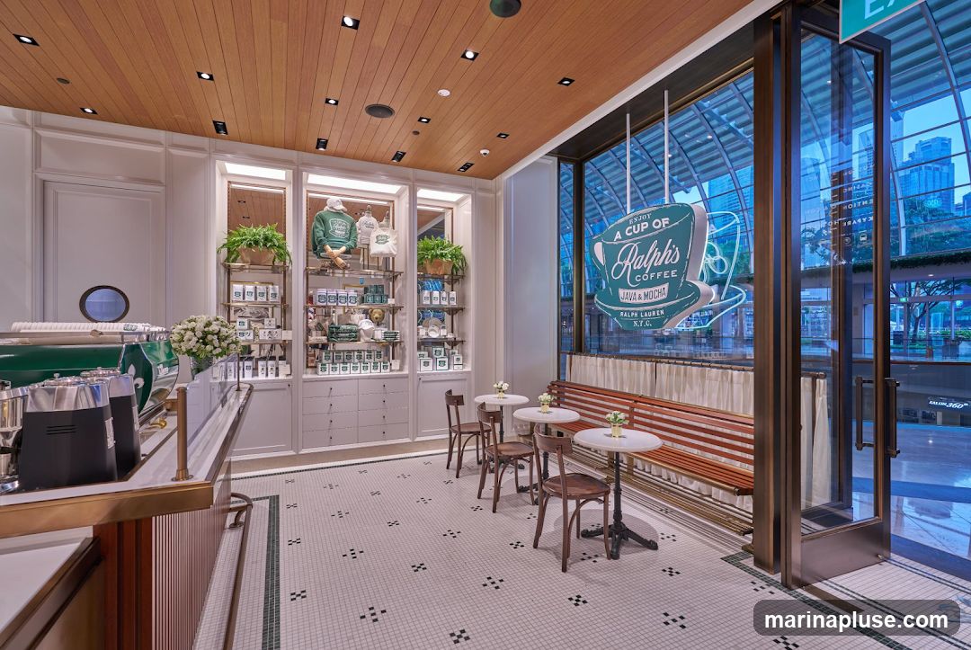 The interior of Ralph's Coffee features a polished aesthetic with a wooden paneled ceiling and tiled floors. A counter with a coffee machine is visible on the left. Display shelves showcase branded merchandise like hoodies and tote bags, alongside packaged coffee and mugs. Seating areas include small round tables with chairs and a long wooden bench. A prominent neon sign in the window reads "Enjoy a cup of Ralph's Coffee Java & Mocha Ralph Lauren N.Y.C.".