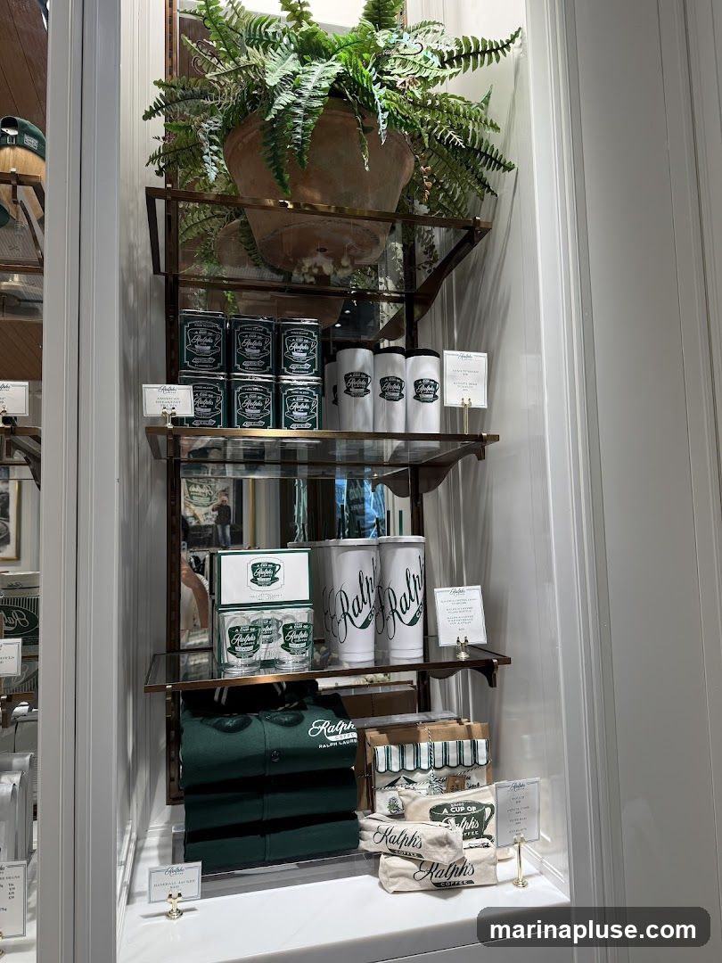 A display window featuring Ralph's Coffee merchandise. On the top shelf, there are several green tins and white cups with the Ralph's logo. The middle shelf holds two boxes of glasses, two large white tumblers, and a small sign. The bottom shelf displays a folded green polo shirt with the Ralph Lauren logo, several bags of coffee beans, and a small sign. A large potted fern sits at the top of the display.