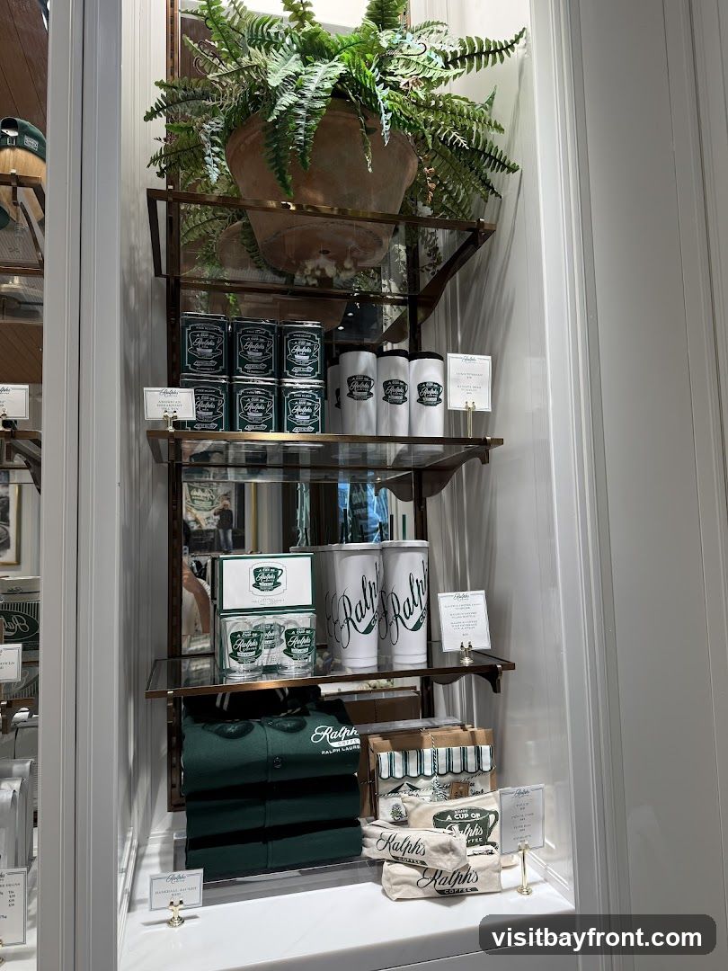 A display window featuring Ralph's Coffee merchandise. On the top shelf, there are several green tins and white cups with the Ralph's logo. The middle shelf holds two boxes of glasses, two large white tumblers, and a small sign. The bottom shelf displays a folded green polo shirt with the Ralph Lauren logo, several bags of coffee beans, and a small sign. A large potted fern sits at the top of the display.