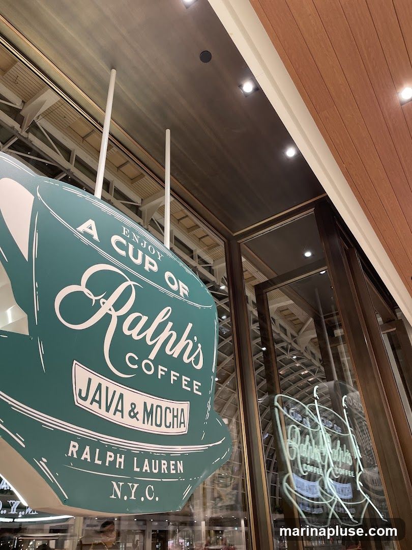 A large, teal-colored sign shaped like a coffee cup, advertising "Ralph's Coffee Java & Mocha" and "Ralph Lauren N.Y.C.". The sign is hanging indoors, possibly in a mall or airport, with reflections of the interior visible in the glass windows. A neon sign with the "Ralph's Coffee" logo is also visible in the reflection.