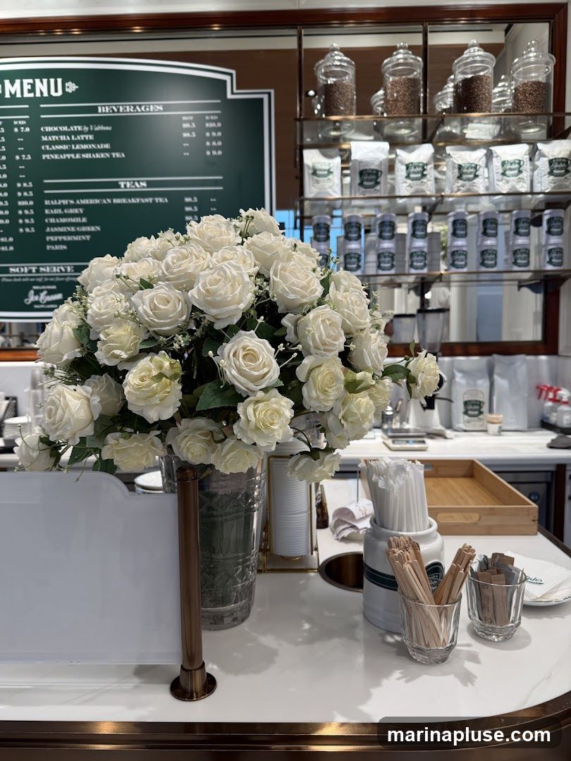 A menu board displays beverages and teas, with a bouquet of white roses in the foreground. Shelves behind the counter are stocked with coffee beans and packaged goods.