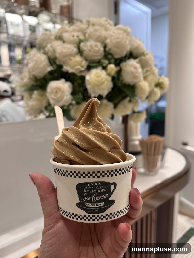 A hand holds a cup of soft-serve ice cream. The cup has a black and white checkered rim and a logo that reads "ENJOY A SCOOP OF DELICIOUS Ice Cream RALPH LAUREN N.Y.C.". In the background, a large bouquet of white roses is visible, along with shelves stocked with various items.