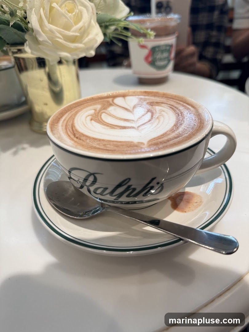 A close-up shot of a latte in a white cup with green trim. The latte has intricate latte art on top. The cup and saucer have the word "Ralph's Coffee" printed on them. A spoon rests on the saucer. In the background, a vase with white roses and greenery is visible, along with another drink in a paper cup.