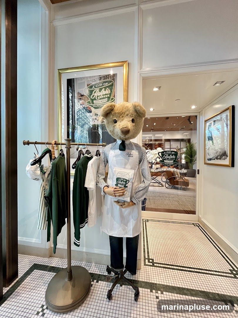A teddy bear dressed as a barista stands in front of a clothing rack displaying green and white apparel. The bear holds a bag of Ralph's Coffee. A framed advertisement for Ralph's Coffee is visible on the wall. The background shows a glimpse into a store interior with seating and more Ralph's Coffee branding.