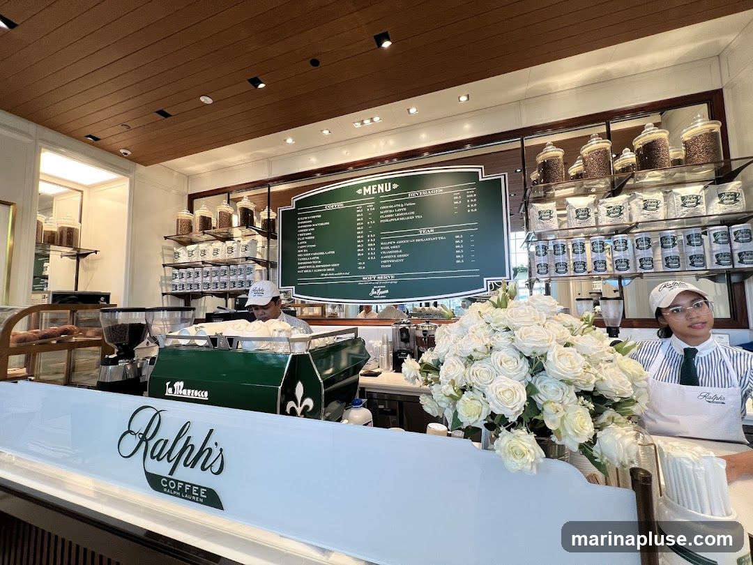 A view of the interior of Ralph's Coffee, showing a counter with a barista, coffee machines, and a menu board. A large bouquet of white roses is prominently displayed in the foreground. Shelves behind the counter are stocked with coffee beans and branded merchandise.