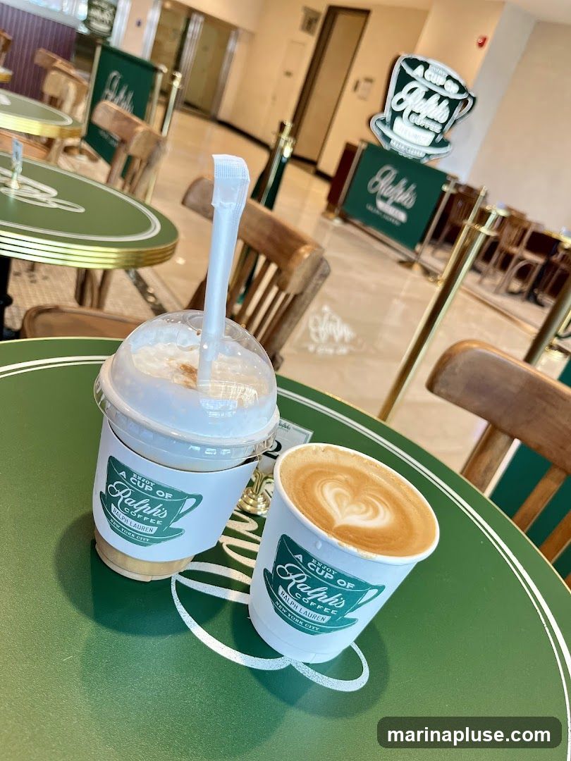 Two coffee drinks, one iced and one hot latte with latte art, sit on a green table with the Ralph's Coffee logo. In the background, the interior of the coffee shop is visible with tables, chairs, and branded signage.
