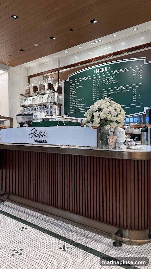 A view of the interior of Ralph's Coffee, showing a counter with a menu board behind it. The counter has a white front with the Ralph's Coffee logo. Behind the counter are shelves with coffee bags and jars, a large bouquet of white roses, and various coffee-making supplies. The menu board lists coffee, beverages, and teas.
