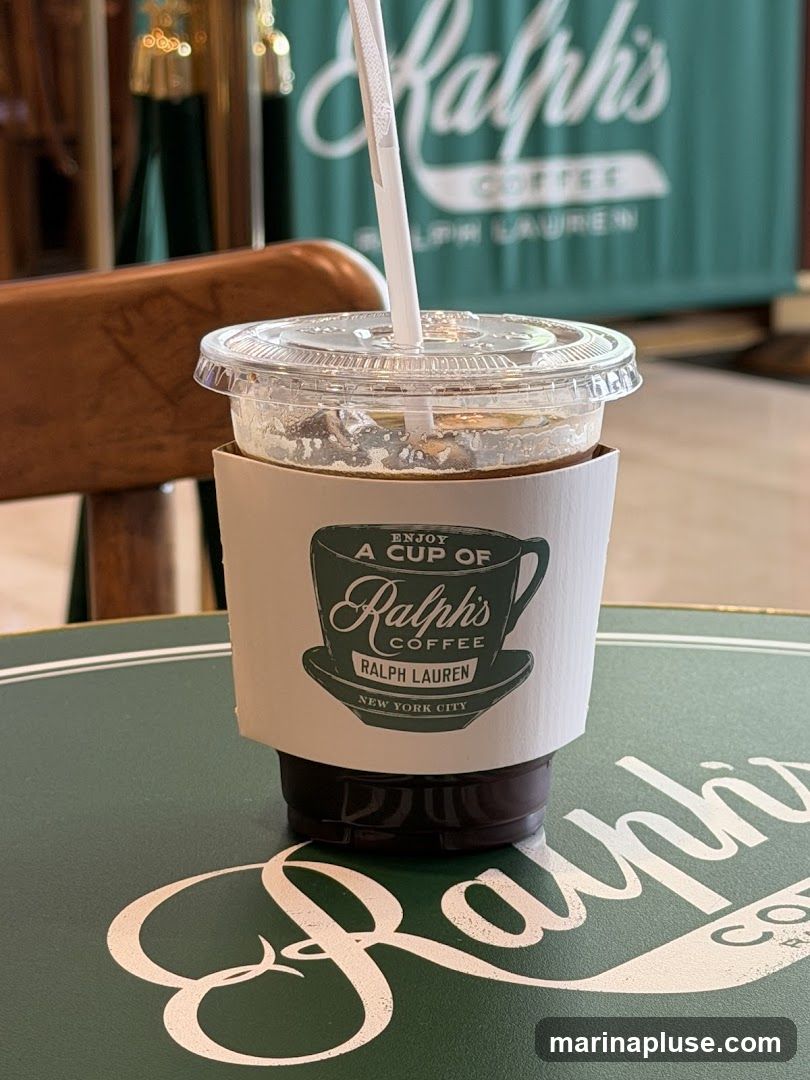 A close-up of an iced coffee drink in a clear plastic cup with a white sleeve. The sleeve has a logo that says "Enjoy a cup of Ralph's Coffee Ralph Lauren New York City" with an illustration of a coffee cup and saucer. The drink is on a green table with a white Ralph's Coffee logo. In the background, a green wall with the Ralph's Coffee logo is visible.