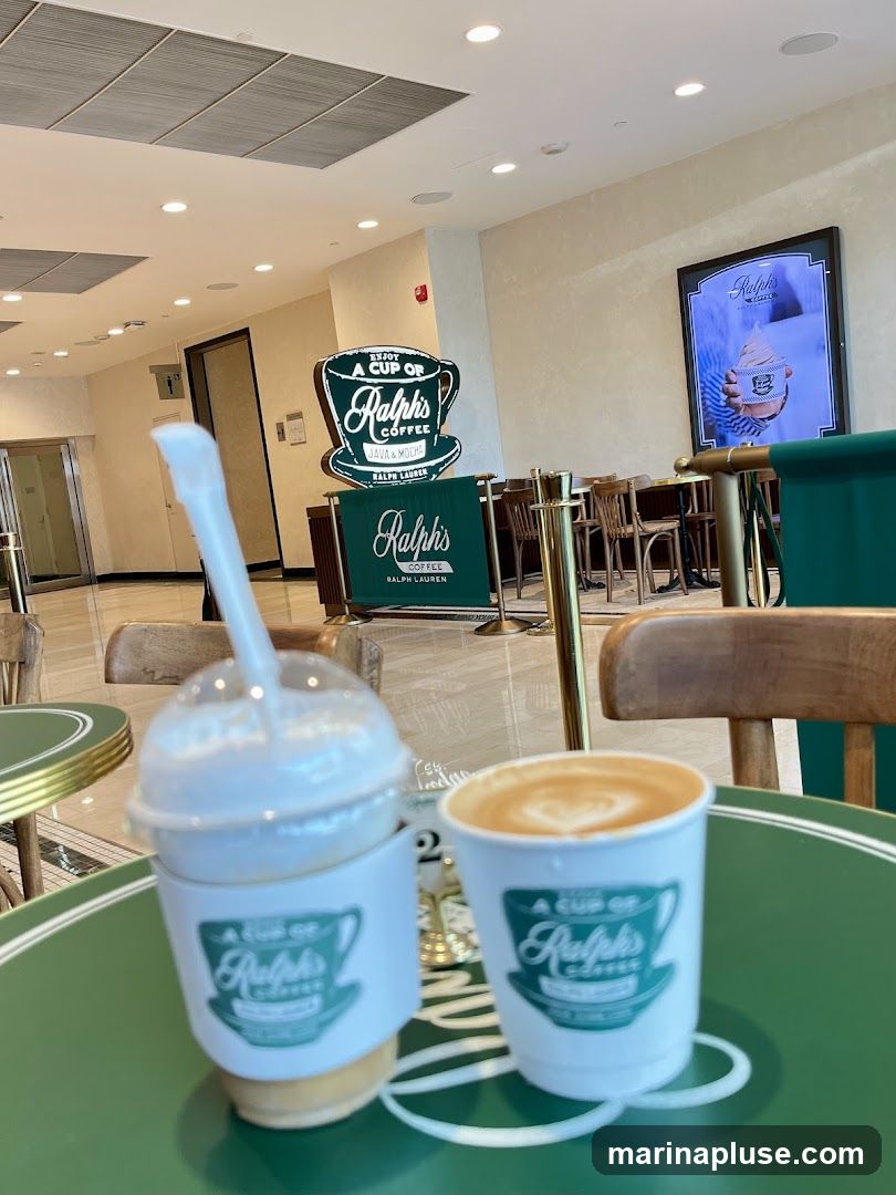 Two coffee drinks are on a green table. One is a latte in a white cup with latte art. The other is a to-go cup with a lid and straw, also in a branded sleeve. In the background, there is a large sign that says "Ralph's Coffee" and "Ralph Lauren". There is also a framed advertisement for Ralph's Coffee.