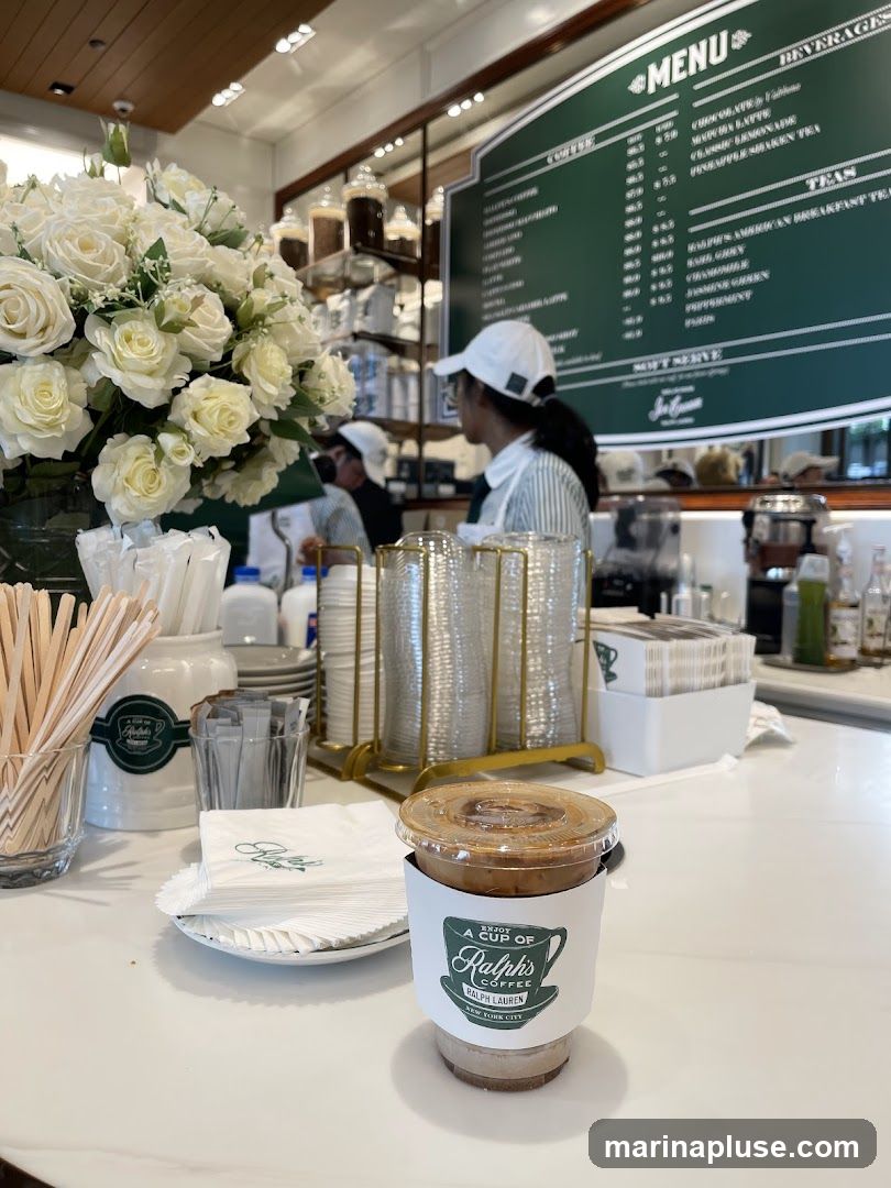 A close-up view of a coffee cup with a Ralph's Coffee sleeve, placed on a white counter. In the background, a menu board displays various beverages and coffee options. A large bouquet of white roses is visible on the left side of the image.