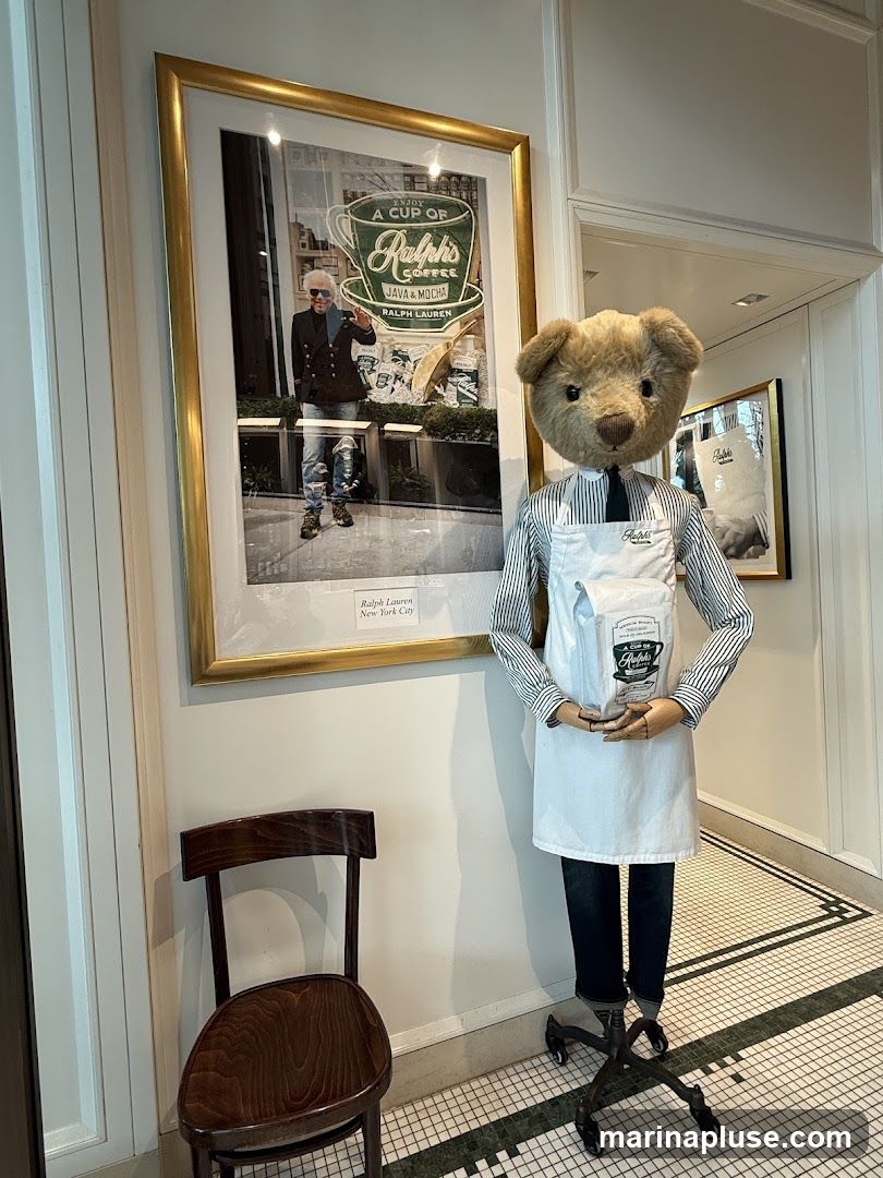 A mannequin dressed as a barista, wearing a striped shirt, white apron, and dark jeans, stands in front of a framed picture. The picture shows Ralph Lauren in a window display with a sign that says "ENJOY A CUP OF Ralph's COFFEE JAVA & MOCHA RALPH LAUREN Ralph Lauren New York City". The mannequin is holding a white bag with the Ralph Lauren logo. A wooden chair is to the left of the mannequin. The floor is tiled with a black and white pattern.