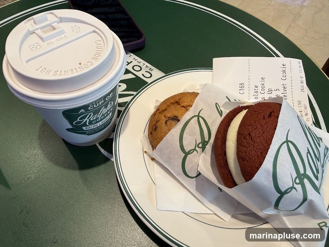 A white paper cup with a white lid sits on a green table. The cup has a logo that says "A CUP OF RALPH'S COFFEE NEW YORK CITY". Next to the cup is a white plate with two cookies. One cookie is a chocolate chip cookie, and the other is a red velvet cookie sandwich with cream filling. A receipt is partially visible under the cookies.